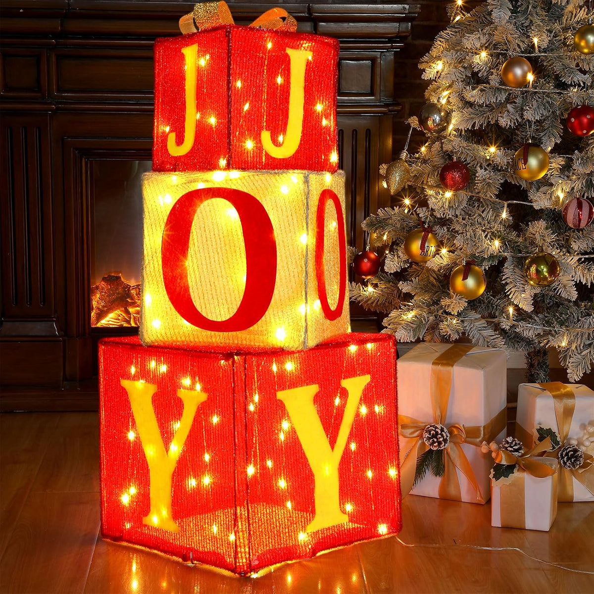 Three stacked lighted red gift boxes with gold letters spelling JOY beside a Christmas tree