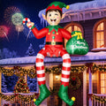 Giant elf inflatable with striped legs sitting on a snowy roof edge holding a candy cane and gift sack